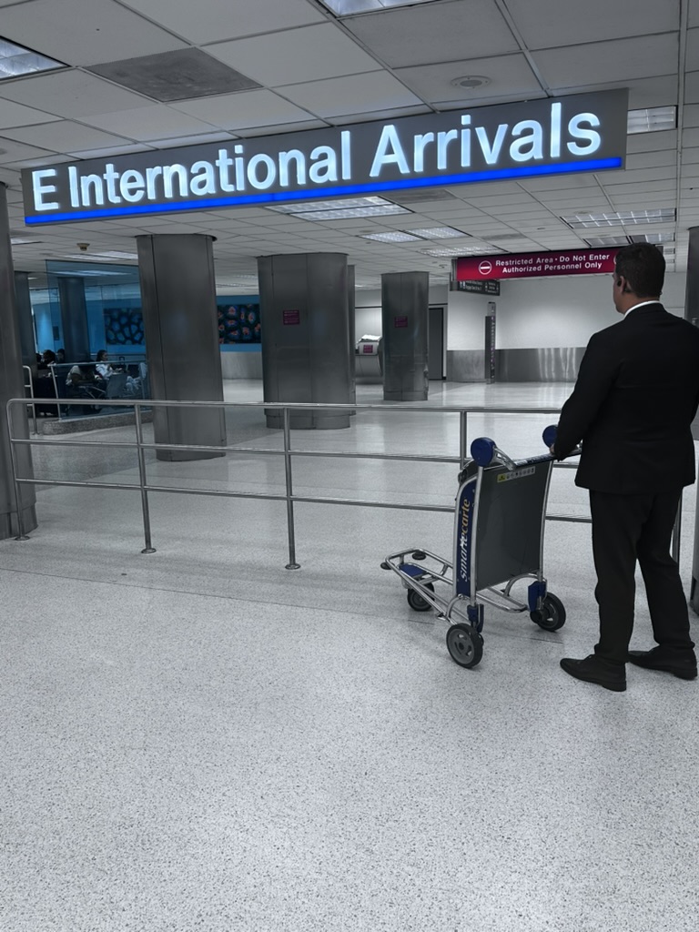 Chauffeur in a dark suit waiting beside a luggage cart beneath the E International Arrivals sign at Miami International Airport.